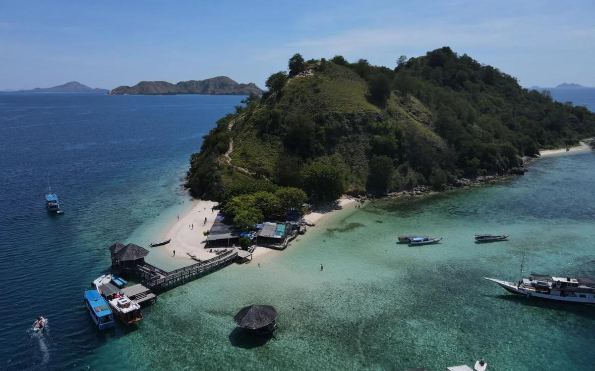 A small green island with a jetty, huts and boats moored in turquoise water