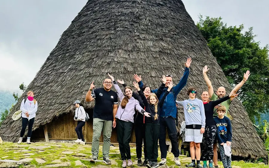 A group of travellers with their arms raised in front of a tall cone-shaped thatched house